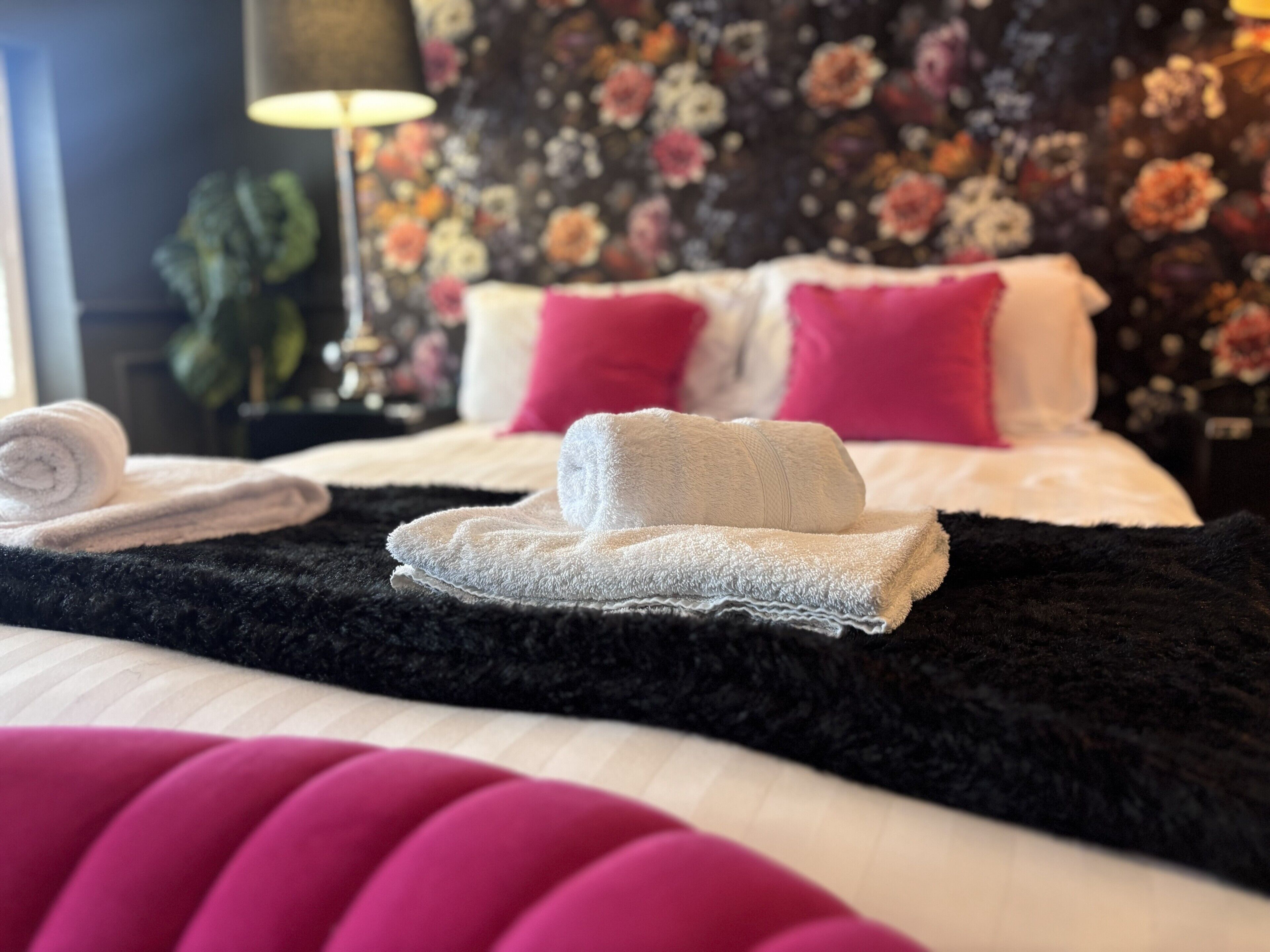 Un lit bien fait, avec des serviettes pliées, une couverture noire et moelleuse, et un oreiller rose.