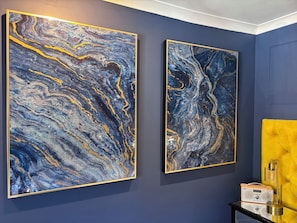 Deux grandes peintures abstraites sur un mur bleu foncĂ©, une tĂȘte de lit jaune Ă motifs et une petite table dâappoint noire avec une radio et un rĂ©cipient en verre.