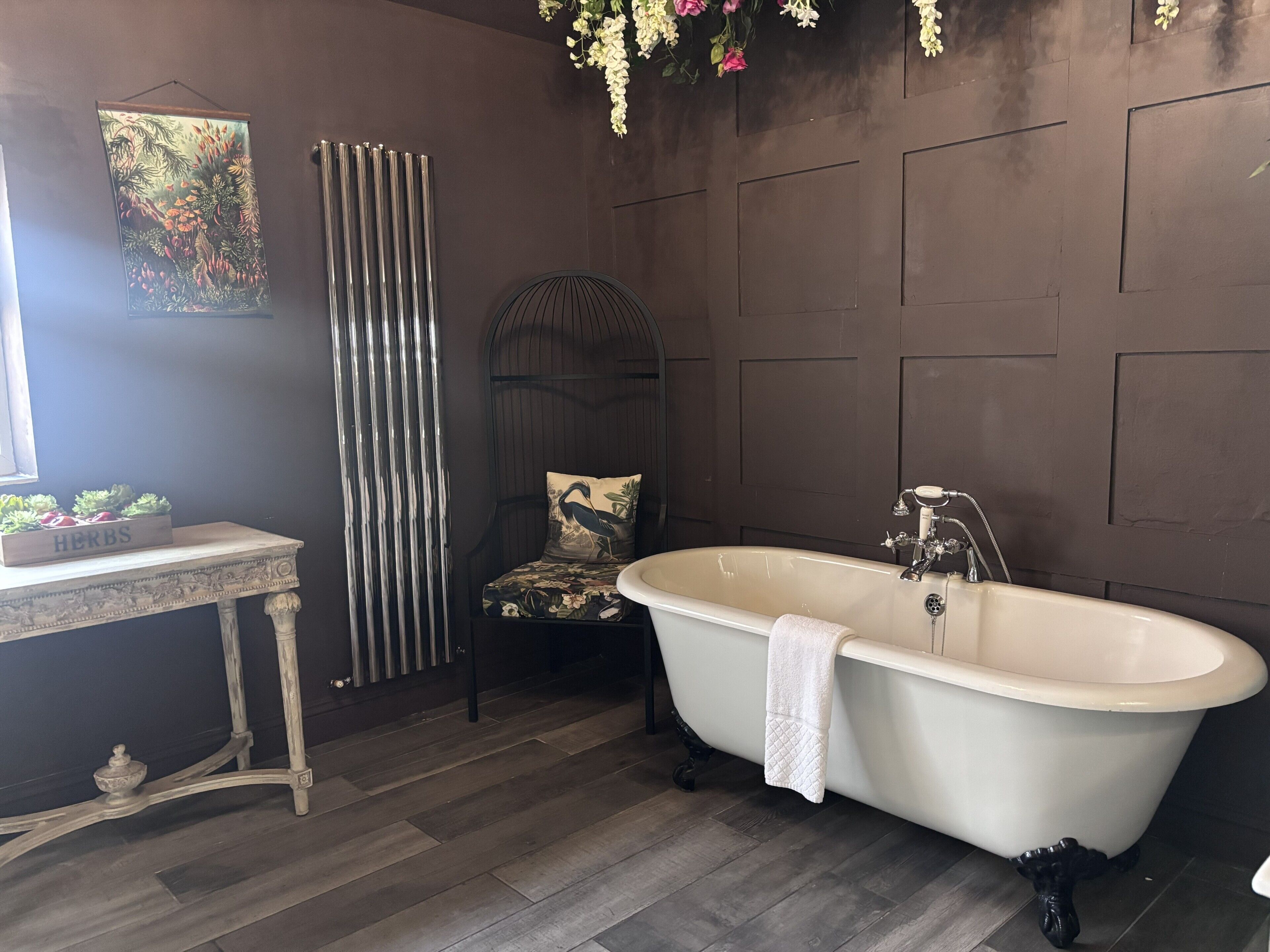 A bathroom with a freestanding bathtub, a radiator, a chair with a floral cushion, and a small table with a herb box.