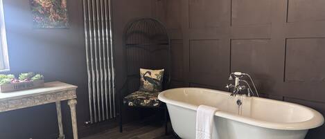 A bathroom with a freestanding bathtub, a radiator, a chair with a floral cushion, and a small table with a herb box.