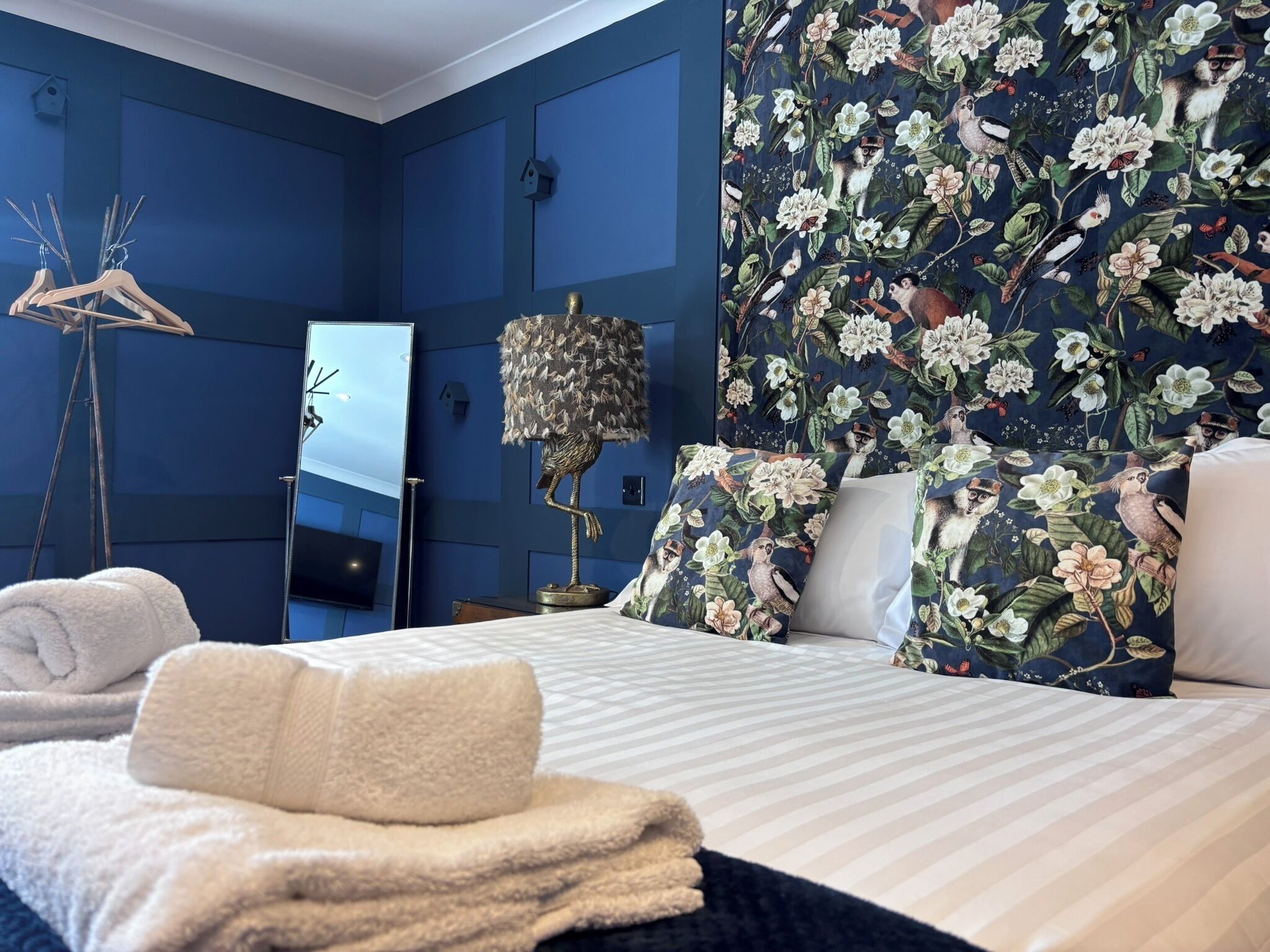 A hotel room with a bed, a mirror, a lamp, and a floral-patterned wall.