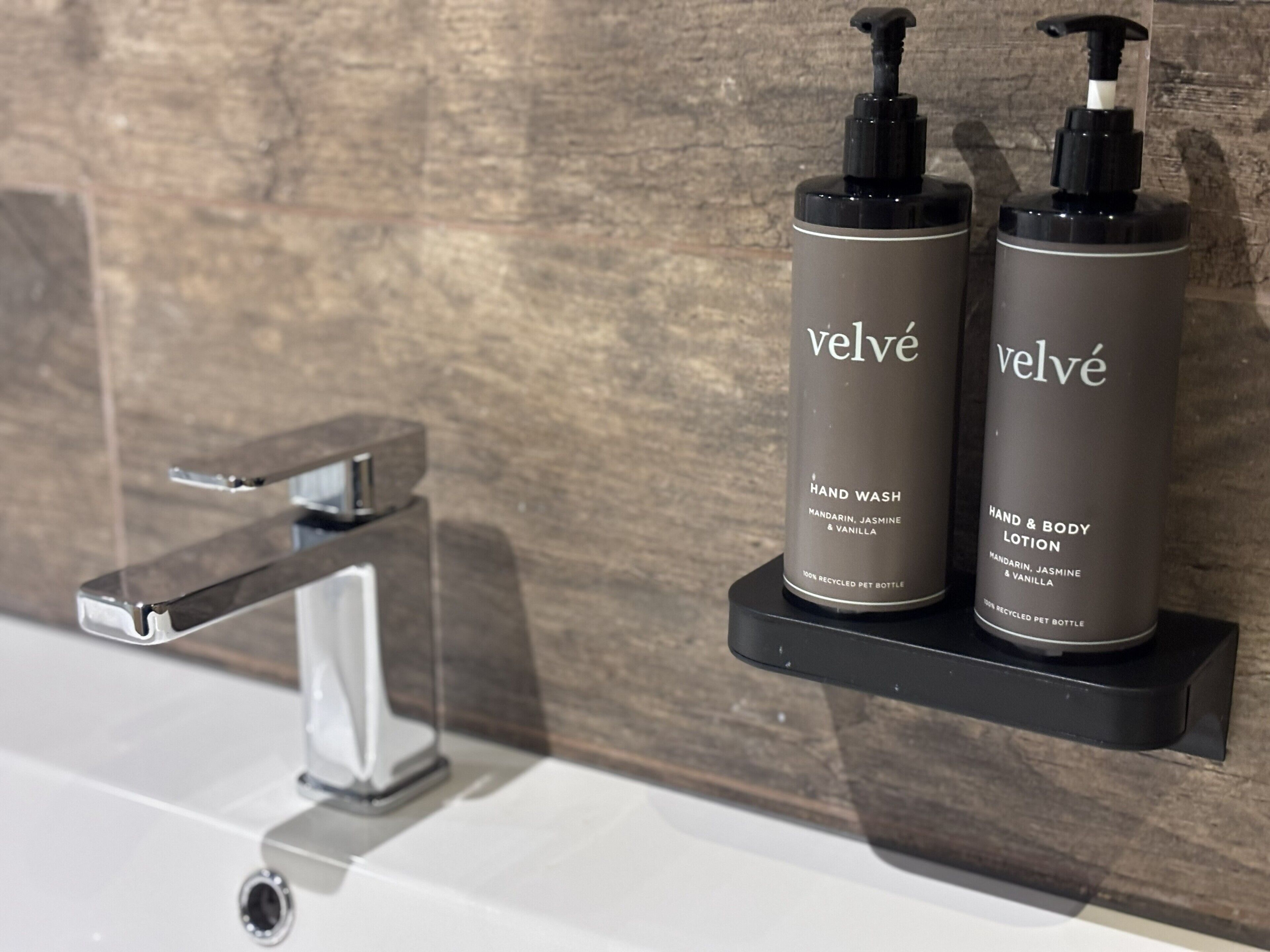 Two bottles of Velvé hand wash and body lotion on a black shelf by a bathroom sink.