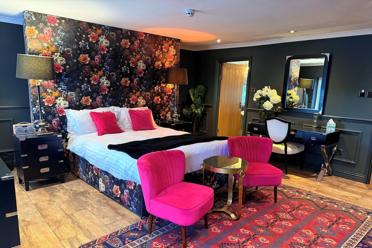 A hotel room with a bed, two chairs, a desk, a mirror, and a floral wall.