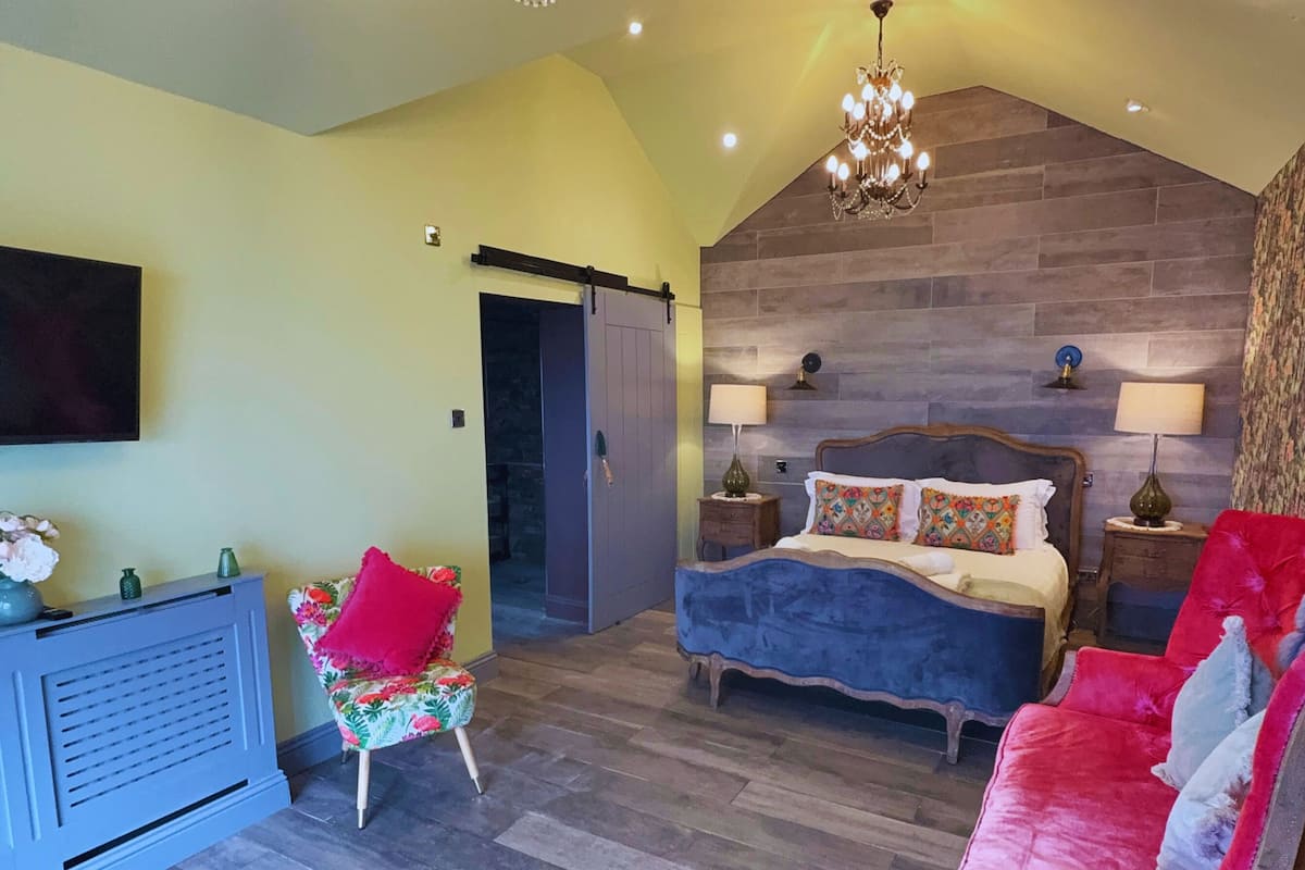 A bedroom with a bed, a chair, a dresser, a chandelier, and a fireplace.