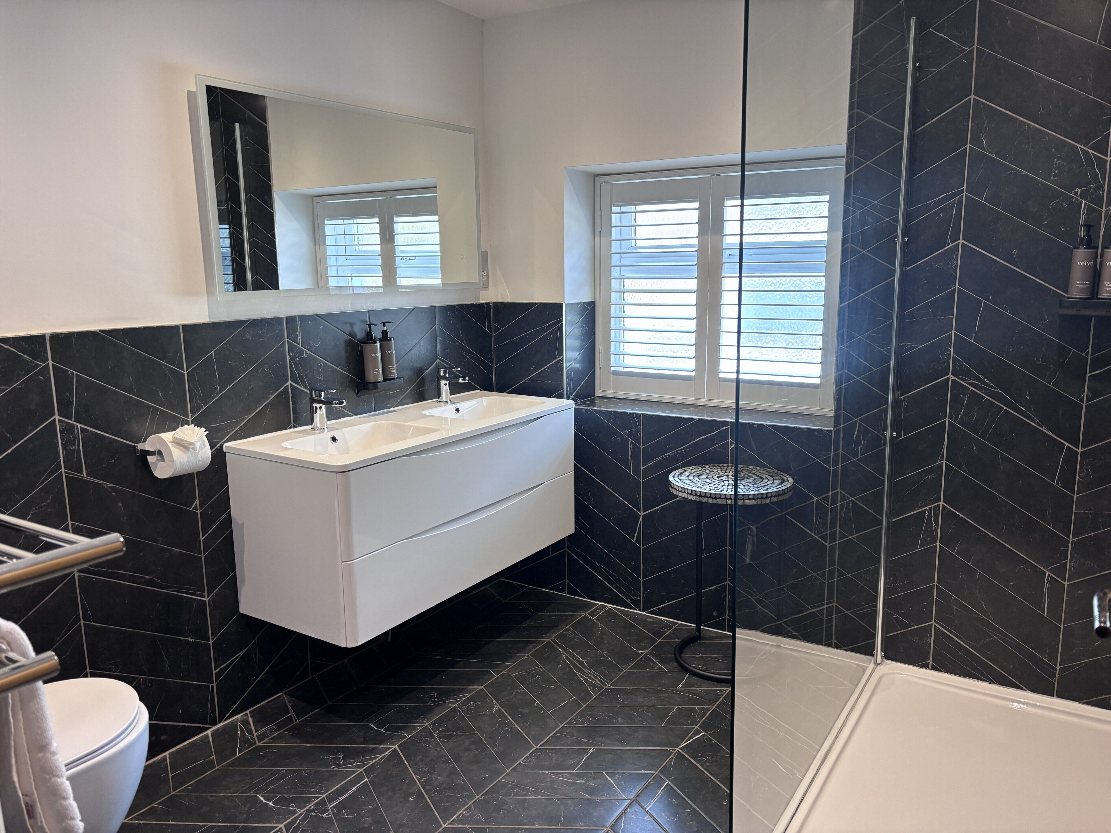 A bathroom with a white vanity, black tiled walls, a window with blinds, and a shower area.