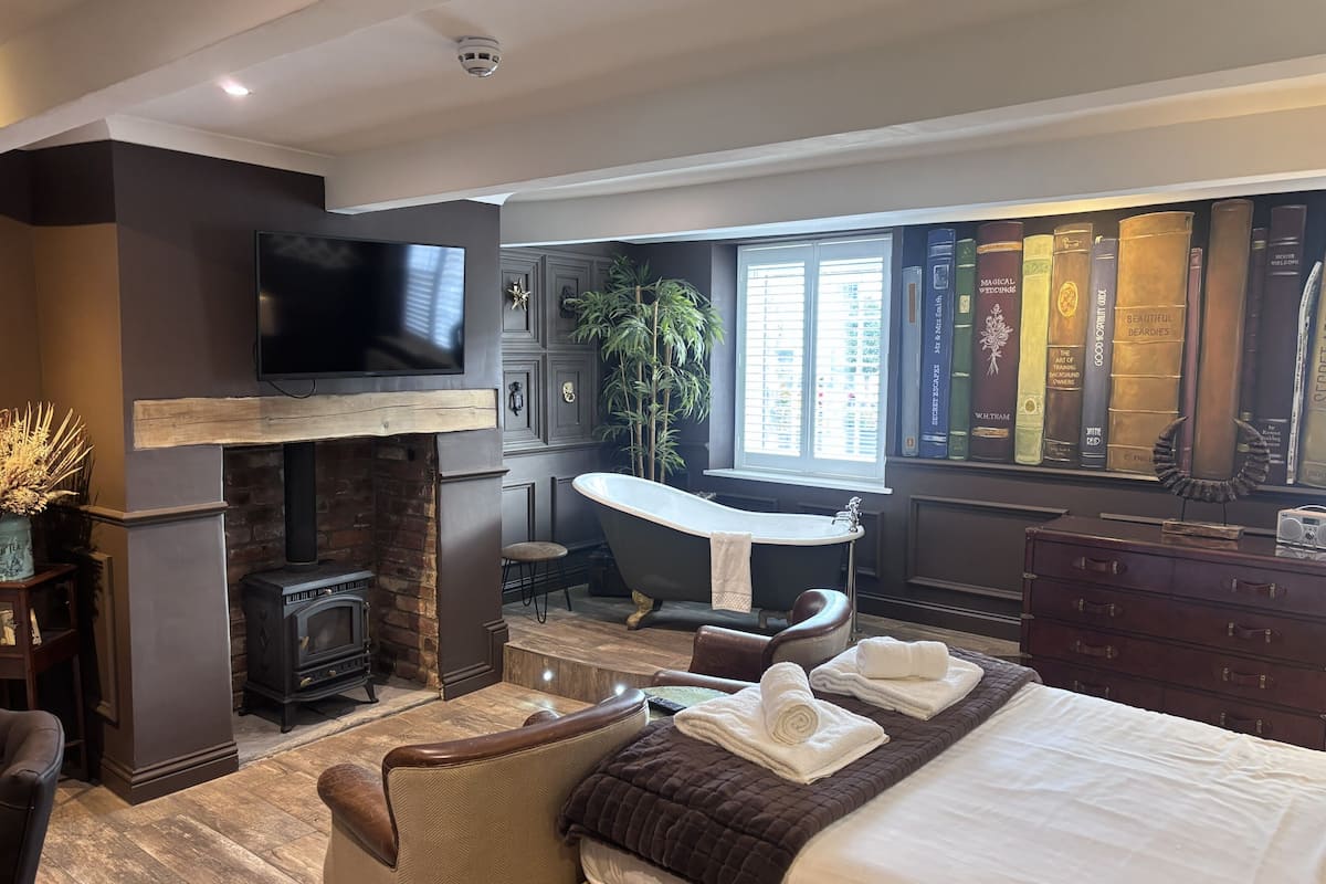 A modern bedroom with a fireplace, a large bed, a bathtub, and a bookshelf.