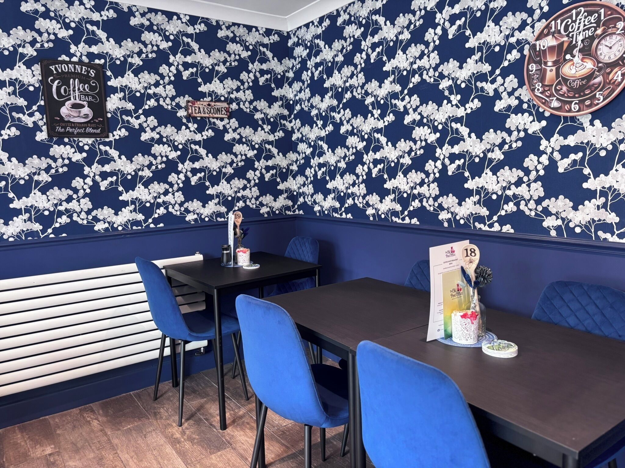 A restaurant interior with blue walls, white floral wallpaper, and blue chairs. There are tables with menus and a coffee machine.
