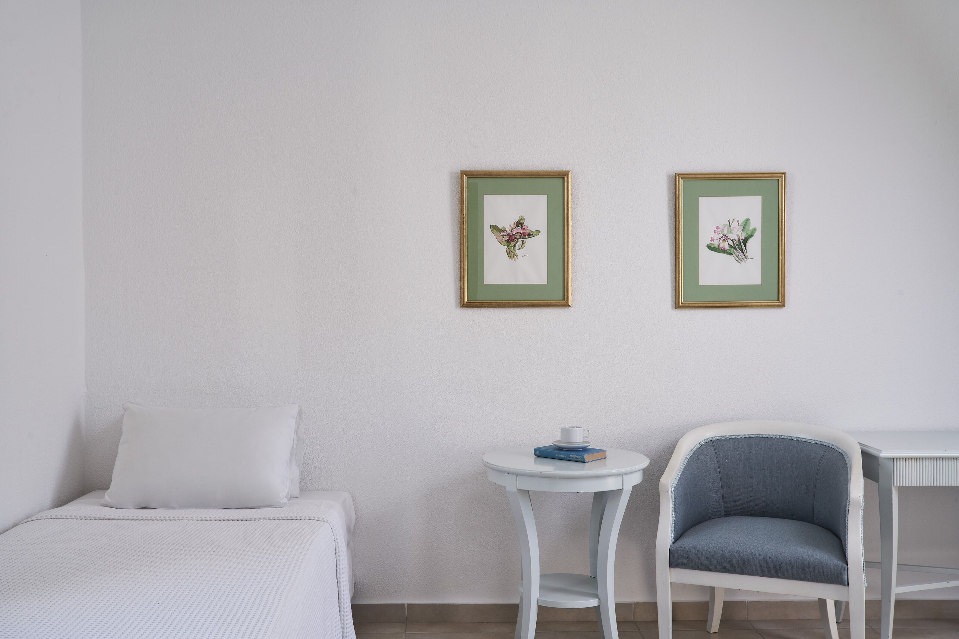 Ein Zimmer mit einem Bett, einem kleinen runden Tisch mit Tasse und Büchern, einem Sessel und zwei gerahmten Pflanzenblättern an der Wand.