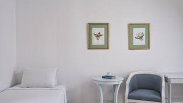 Ein Zimmer mit einem Bett, einem kleinen runden Tisch mit Tasse und Büchern, einem Sessel und zwei gerahmten Pflanzenblättern an der Wand.