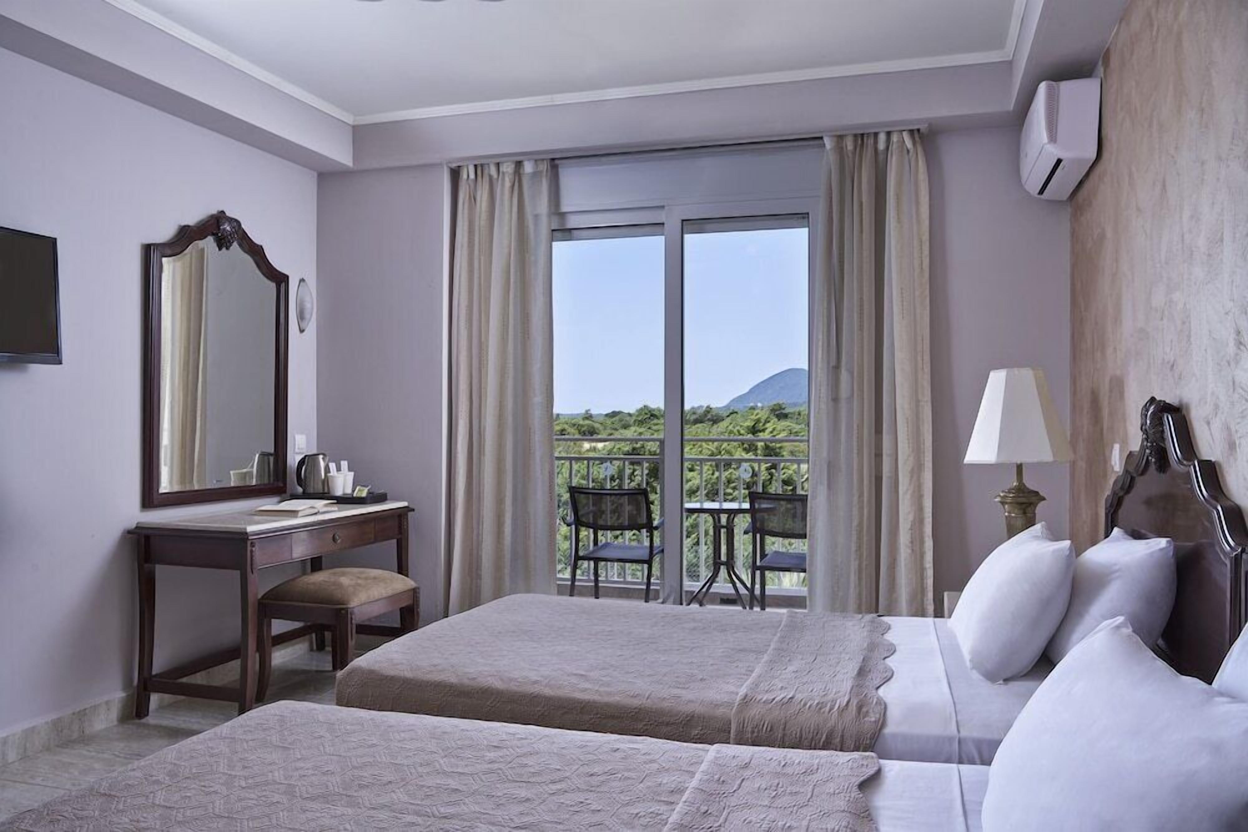 Habitación de hotel con dos camas, un escritorio, una silla, un espejo grande y un balcón con vistas a la vegetación y una montaña.