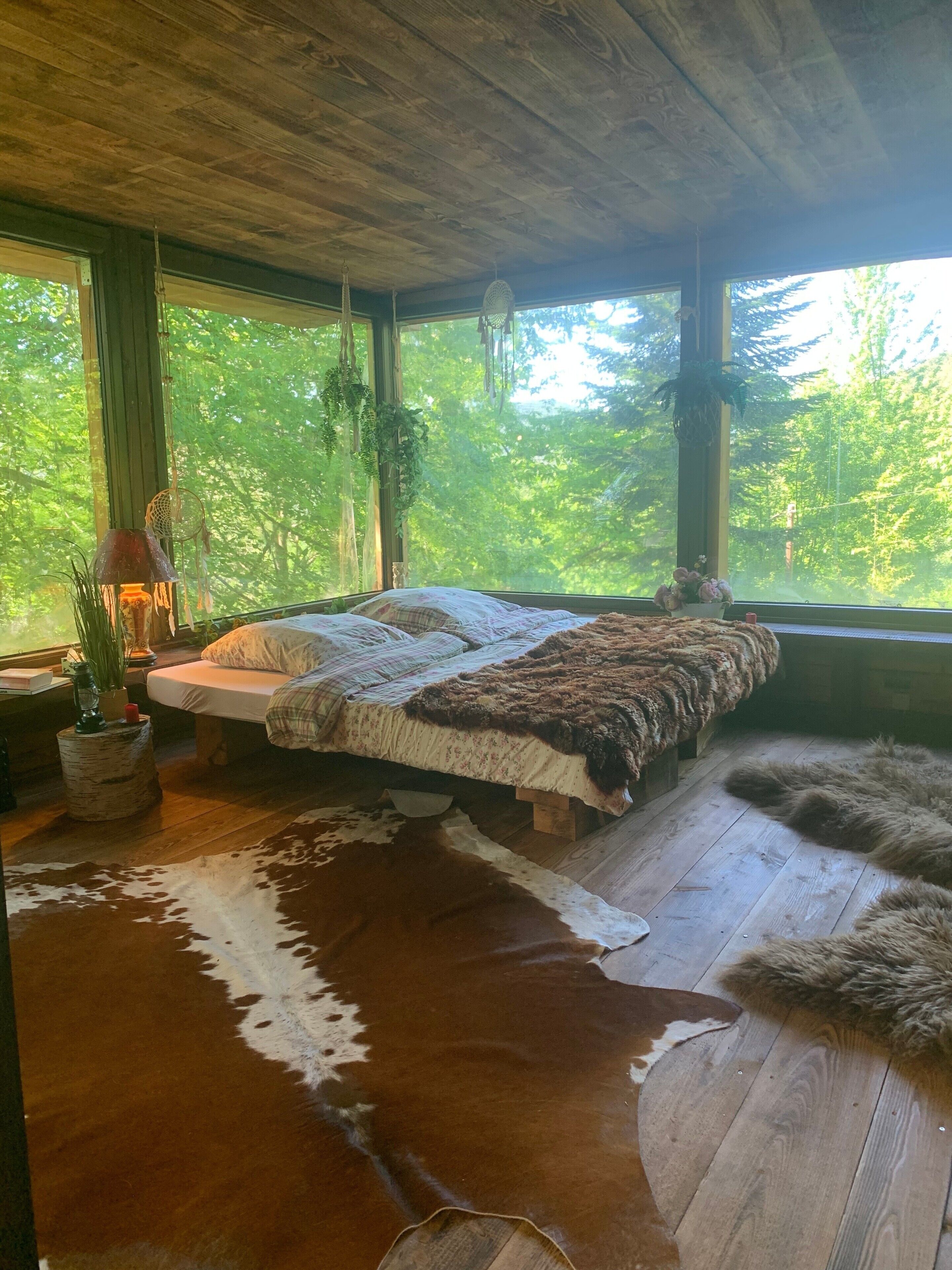 A rustic room with a bed, wooden floor, and large windows overlooking greenery.