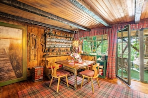Interior rústico de madera con piso estampado en tartán, una mesa de madera con sillas y una ventana con vista a follaje.