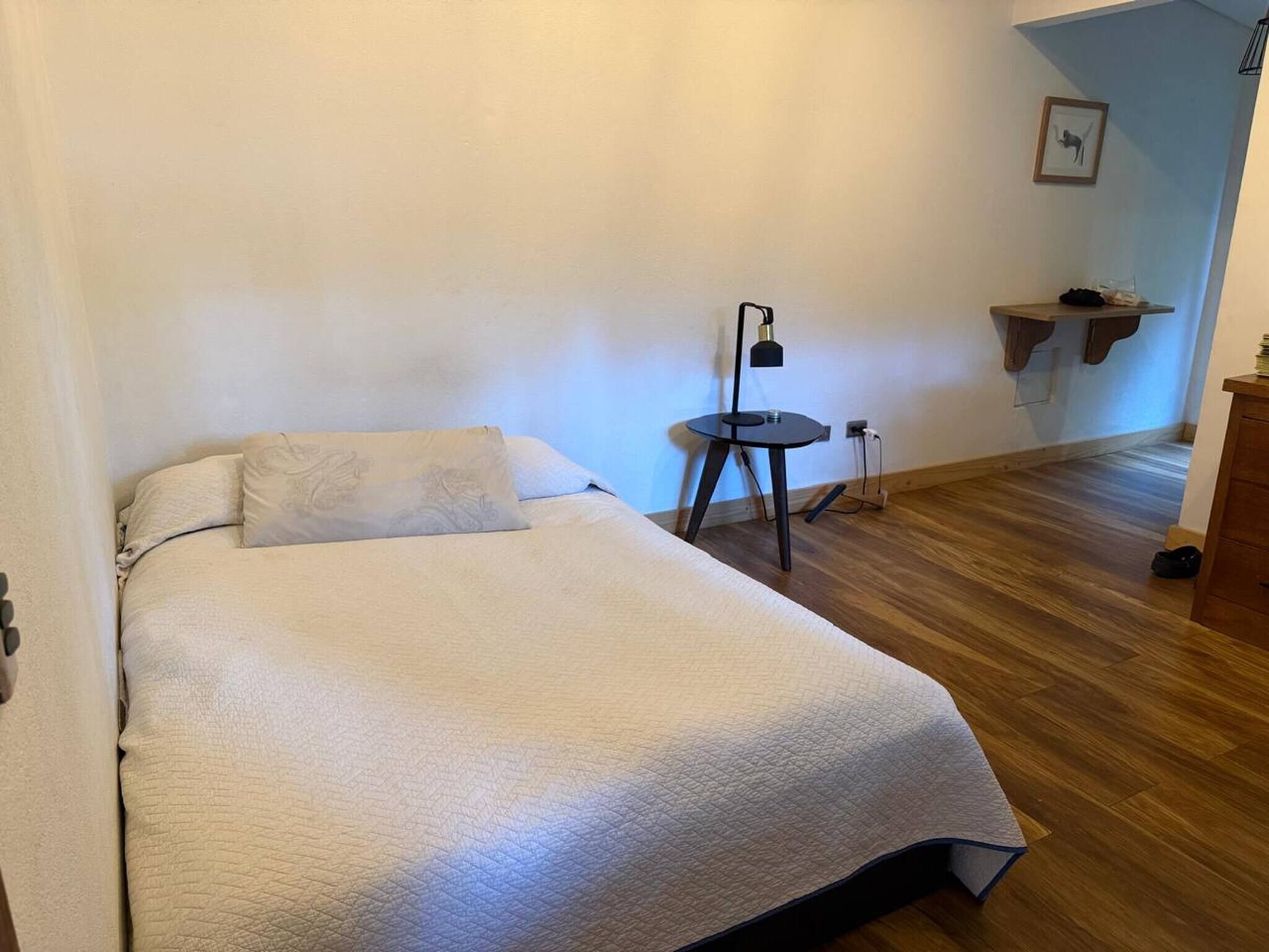 Une chambre à coucher avec un lit, une petite table avec une lampe, un plancher en bois et une étagère fixée au mur.