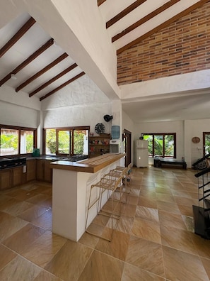 A spacious kitchen with a central island, wooden beams, large windows, and a brick wall.