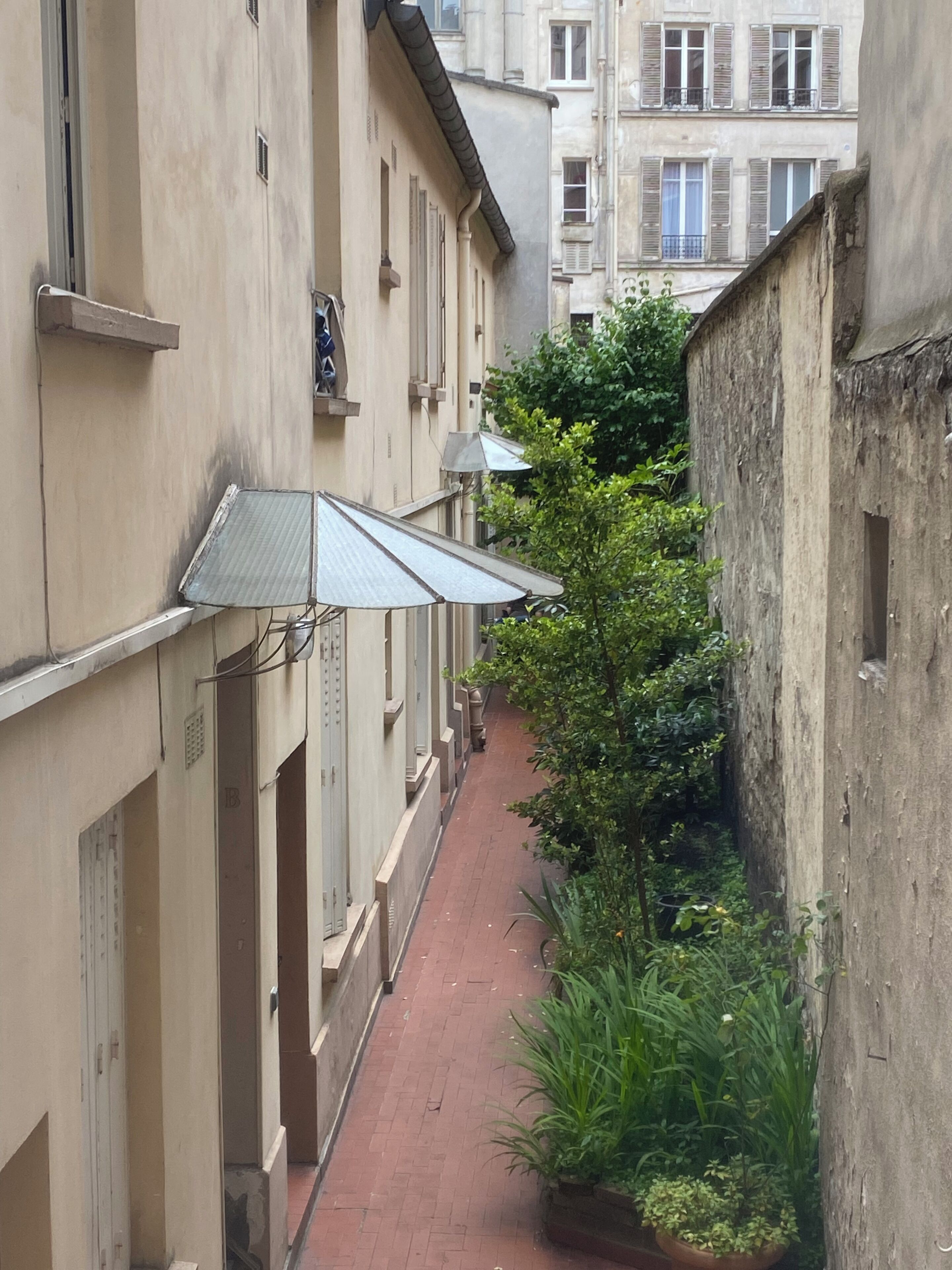 A narrow alleyway between buildings with a red brick path, metal awnings, and greenery.
