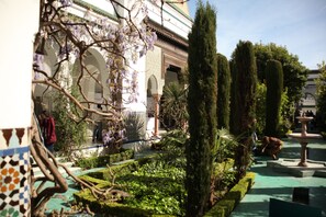 Une cour avec une fontaine, des haies taillées et un bâtiment orné d’arcades.