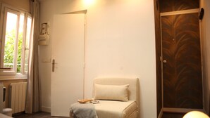 A room with a door, a radiator, a window with curtains, a chair with a cushion, and a door with a palm leaf pattern. - Bohemian & Historic Paris Panthéon - Mosquée (Paris)