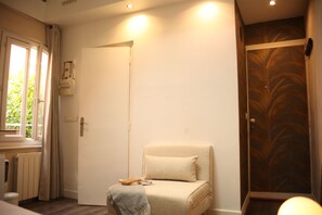 Une pièce avec une porte, un radiateur, une fenêtre avec des rideaux, une chaise avec un coussin et une porte ornée d’un motif de feuilles de palmier.