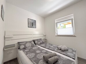 Ein Einzelbett mit Kopfteil, ein kleines Fenster mit Jalousien, ein Wandbild und eine Steckdose.