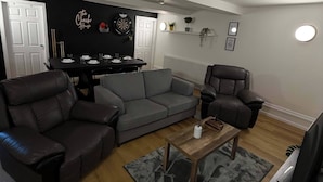 Un salon avec un canapé gris, deux fauteuils, une table basse, un téléviseur et un jeu de fléchettes accroché au mur.