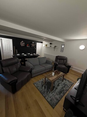 A living room with a grey sofa, two armchairs, a coffee table, a TV, and a dartboard on the wall. - The Coach House, Huddersfield (West Yorkshire)