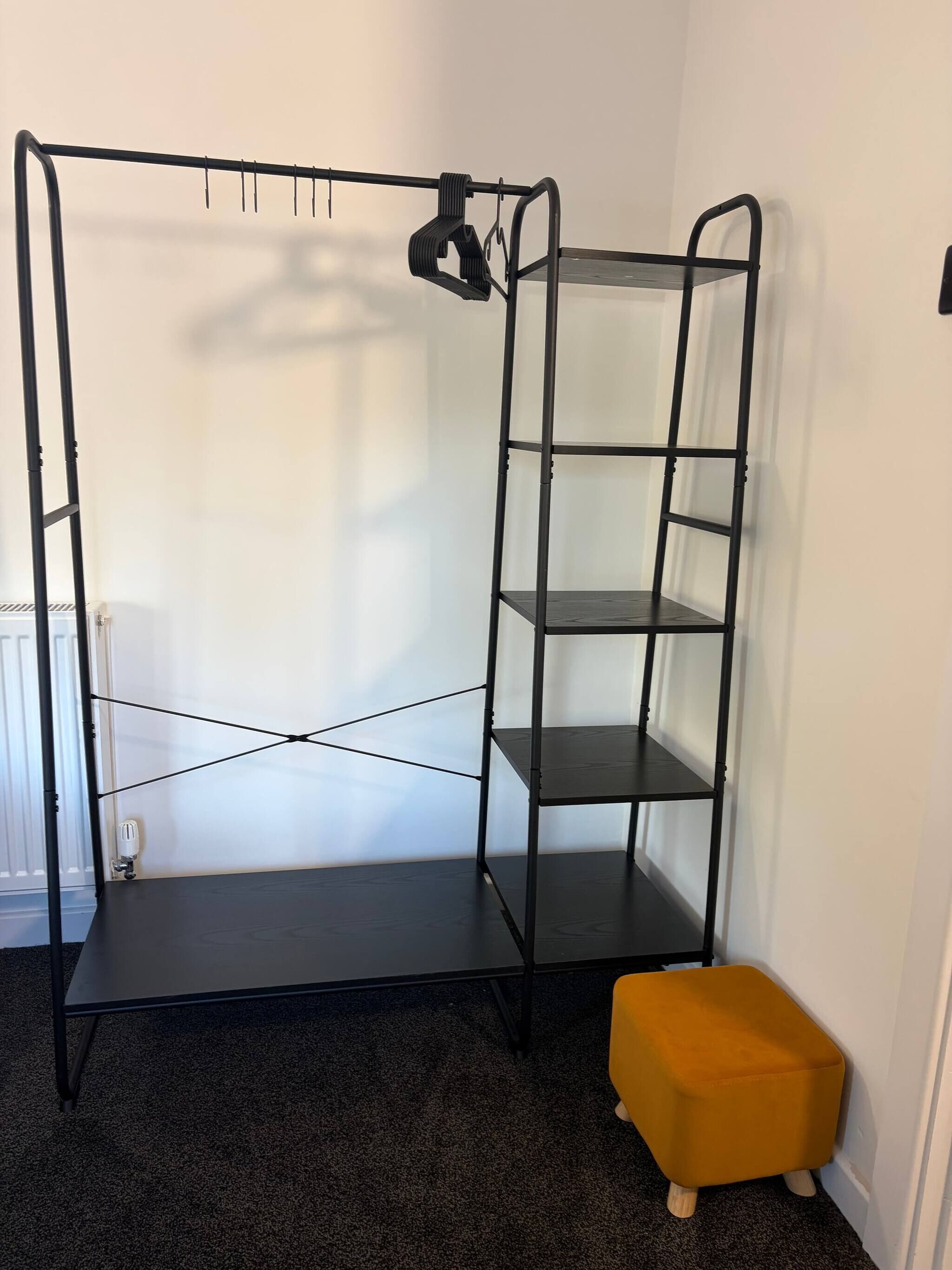 A room with a metal shelving unit, a radiator, and a small orange ottoman.