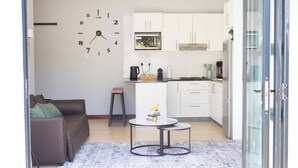 Un espacio habitable compacto con kitchenette, un sofá, una mesita redonda y un reloj de pared.