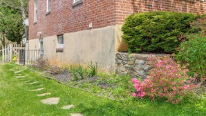 Un edificio de ladrillo con un muro de contención de piedra, un jardín cuidado con piedras para caminar y una cerca blanca de piquetes.