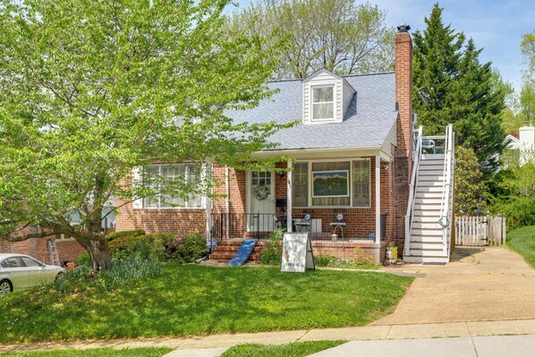A two-story house with a front porch, a visible garage door, and a small front yard. - Gateway to Downtown Baltimore! Cozy & Private Unit (Baltimore)