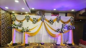 A stage with draped curtains and floral arrangements, a patterned carpet, and a ceiling fan. - Hotel Om Shivay  (Raebareli)