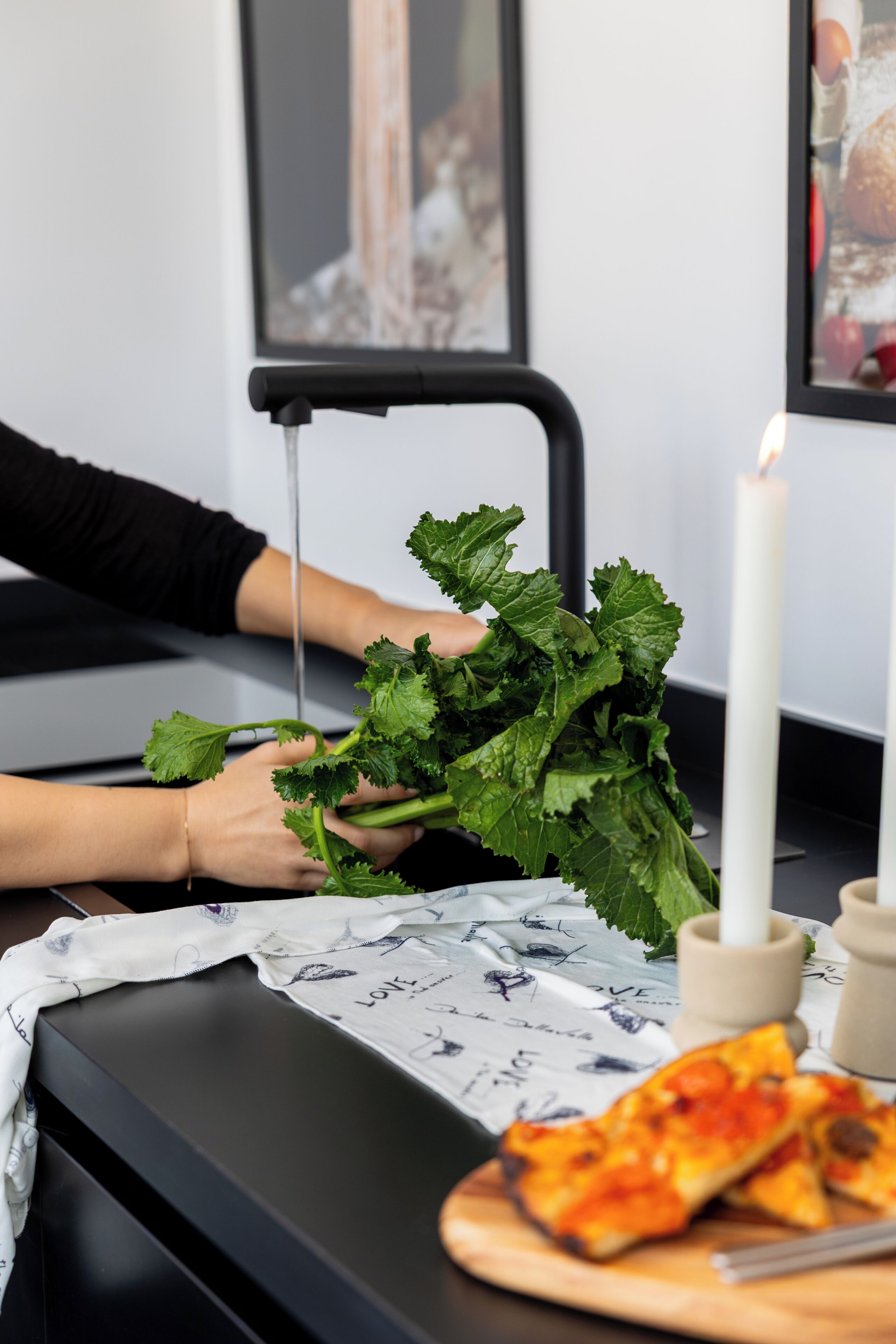 Un comptoir de cuisine avec une planche à découper, un couteau, une assiette contenant des parts de pizza, une bougie blanche et une personne tenant des feuilles de salade.