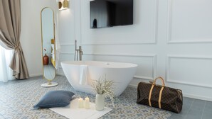 Un baño moderno con una bañera independiente, un espejo grande, un televisor de pantalla plana y un bolso Louis Vuitton en el suelo.