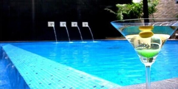 A clear glass of a green cocktail with a garnish, placed on a poolside with a blue pool and a stone wall in the background. - Inventree Hotel Bangalore (BENGALURU)