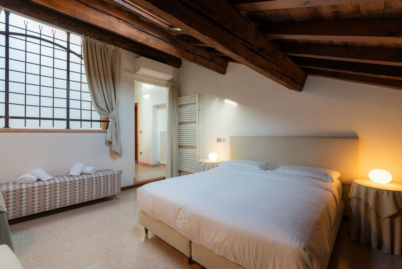 Ein Schlafzimmer mit einem großen Bett, einem Nachttisch mit Lampe, einer Bank mit Handtüchern und einem Fenster mit Vorhängen.