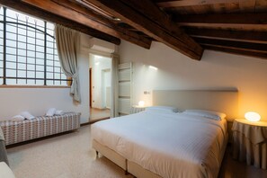 A bedroom with a large bed, a bedside table with a lamp, a bench with towels, and a window with curtains. - Elegant estate overlooking Piazza Erbe (Verona)