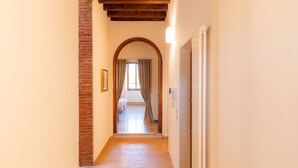 Un pasillo con pared de ladrillo, vigas de madera en el techo y una puerta que da a una habitación con cama y cortinas.