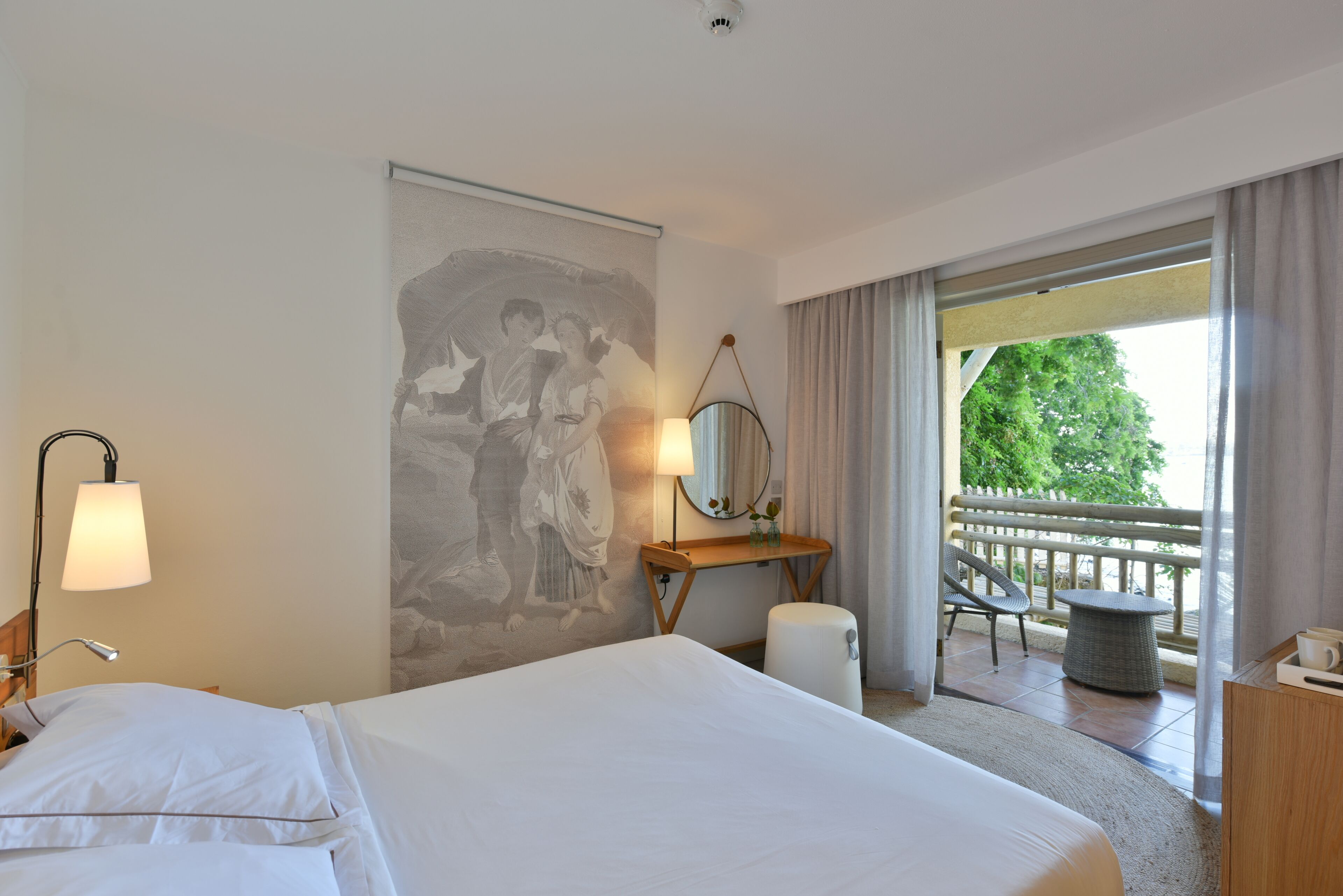 A hotel room with a bed, a desk, a chair, a window with curtains, and a balcony with a view of greenery.