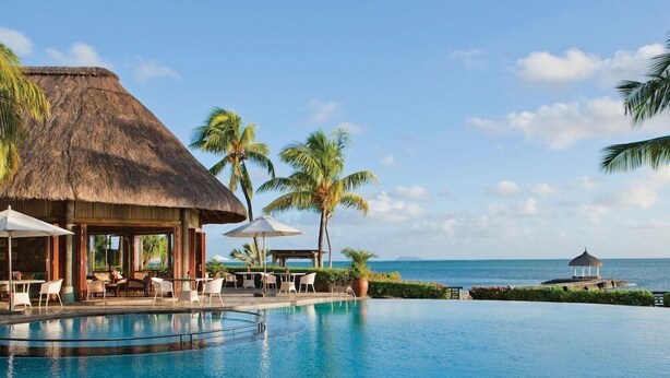 A resort pool with lounge chairs, a thatched-roof building, palm trees, and a view of the ocean. - Veranda Paul et Virginie Hotel and Spa - Adults Only (Grand Gaube)