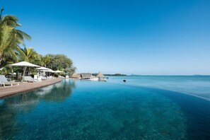 A clear blue infinity pool with lounge chairs and umbrellas, surrounded by palm trees and a thatched-roof structure.