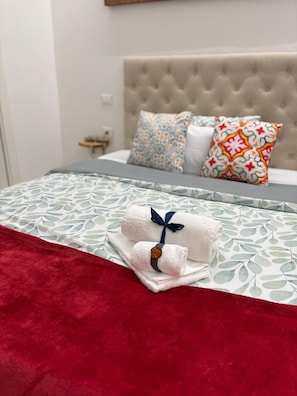 Una cama bien hecha con una colcha roja, una toalla blanca con un lazo azul y almohadones estampados.