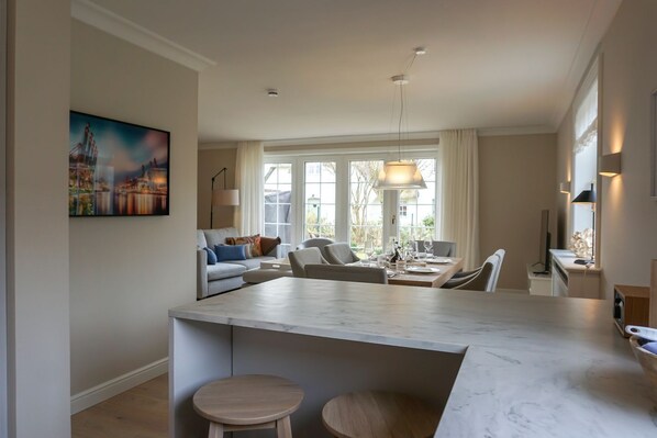 A modern kitchen with a marble island, a dining area with a table set for four, and a living room with a sofa and chairs.