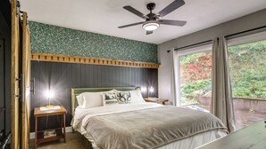 Un dormitorio con cama, mesitas de noche, ventilador de techo, ventana con cortinas y vista a zonas verdes.