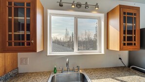 Uma cozinha com bancadas de granito, pia de aço inoxidável, armários de madeira, janela com vista para árvores e luminária no teto.
