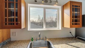 A kitchen with granite countertops, a stainless steel sink, wooden cabinets, a window with a view of trees, and a ceiling light fixture.