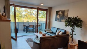 En stue med sofa, spisebord og stole. Der er en skydedør, der fører til et udendørs område med et bord og stole. Et billede hænger på væggen, og en potteplante står på gulvet.