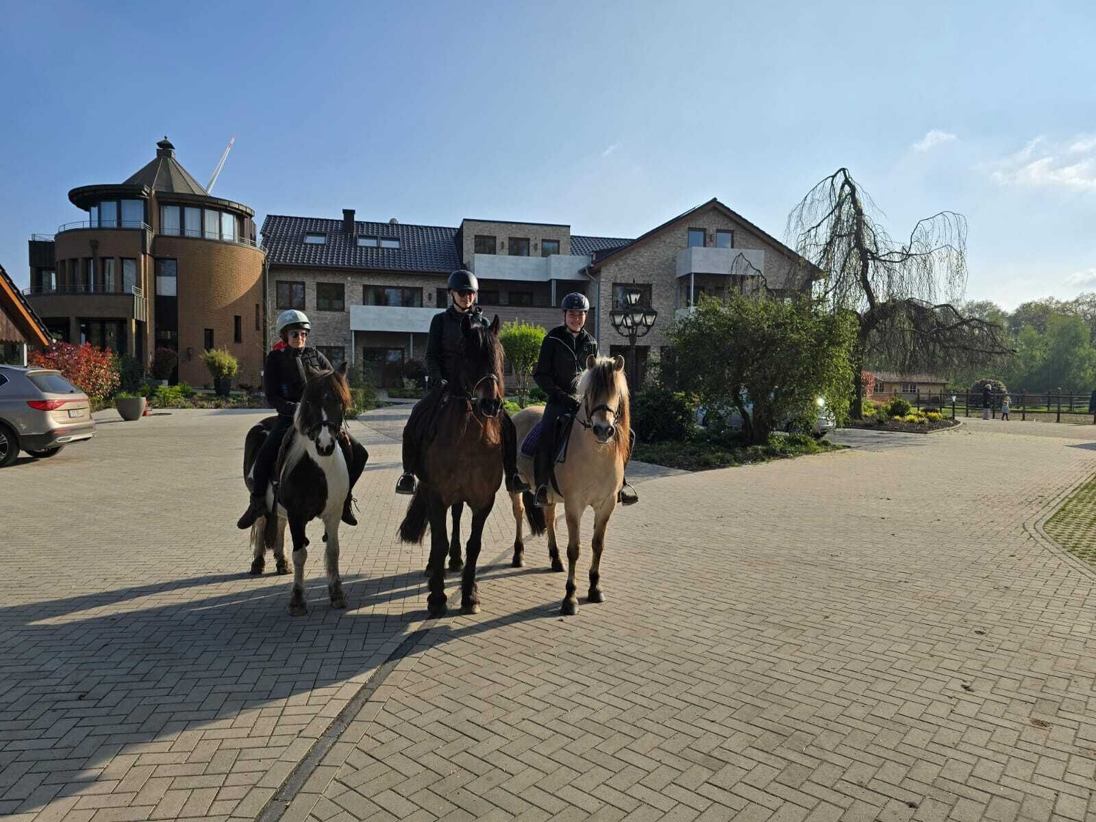 Tre personer rider på hästar på en asfalterad yta, med byggnader i bakgrunden.