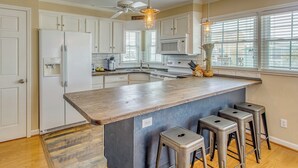 Una cucina con mobili bianchi, un'isola in legno, sgabelli da bar, una ventola a soffitto e una grande finestra.