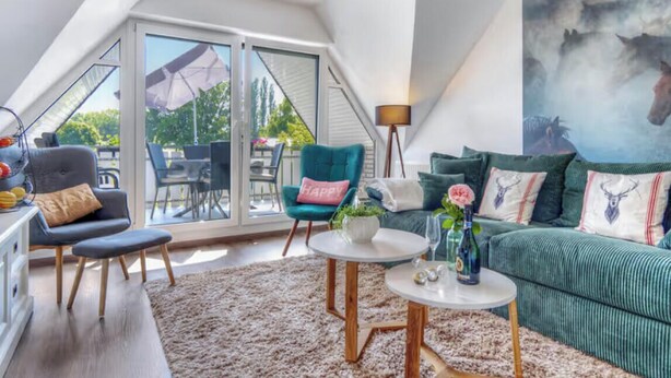 Et moderne loftværelse med skråvæg, stort vindue, grøn sofa, lænestol i teal farve og et billede af heste.