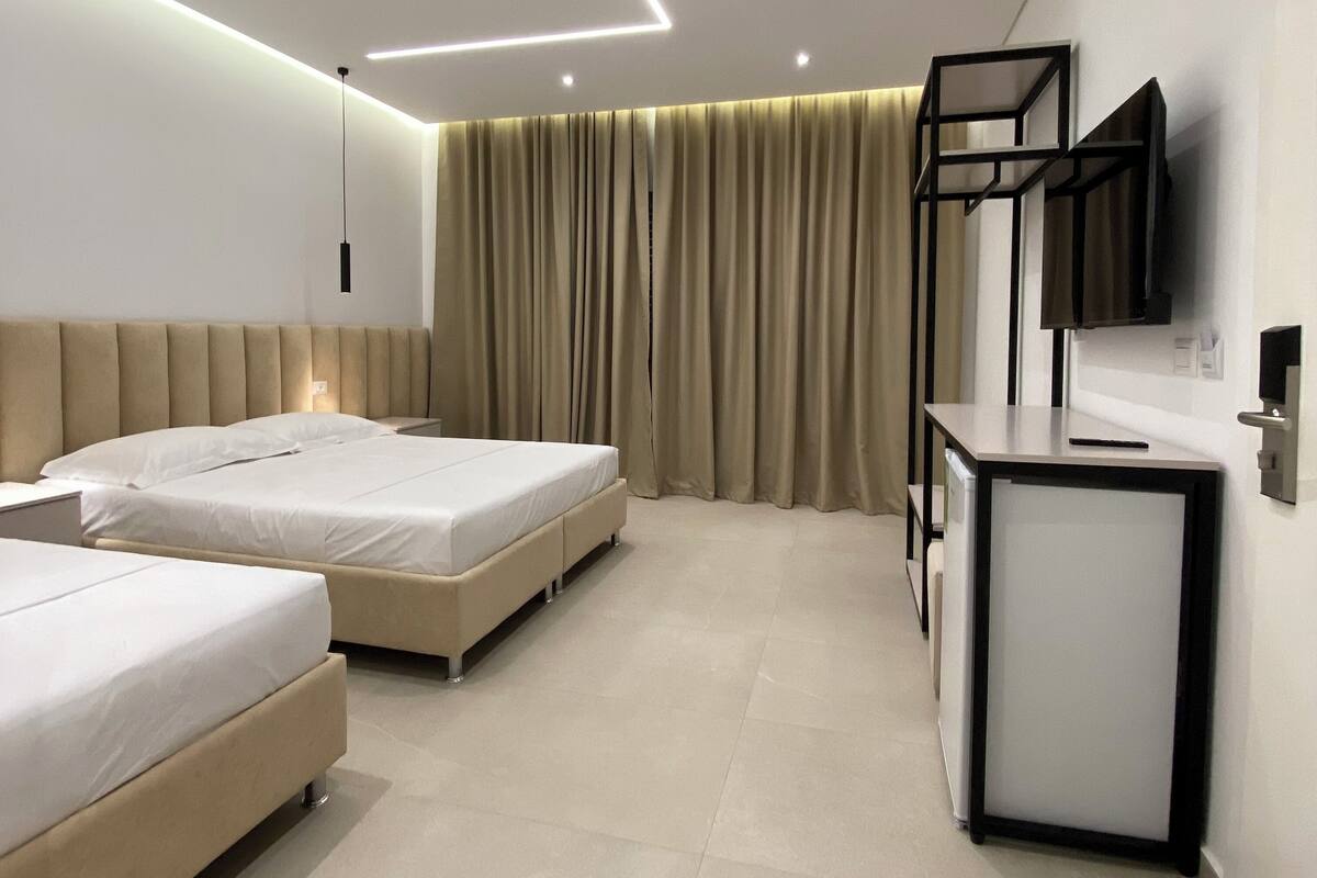 A modern hotel room with two beds, a desk, a flat-screen TV, and a large window with curtains.