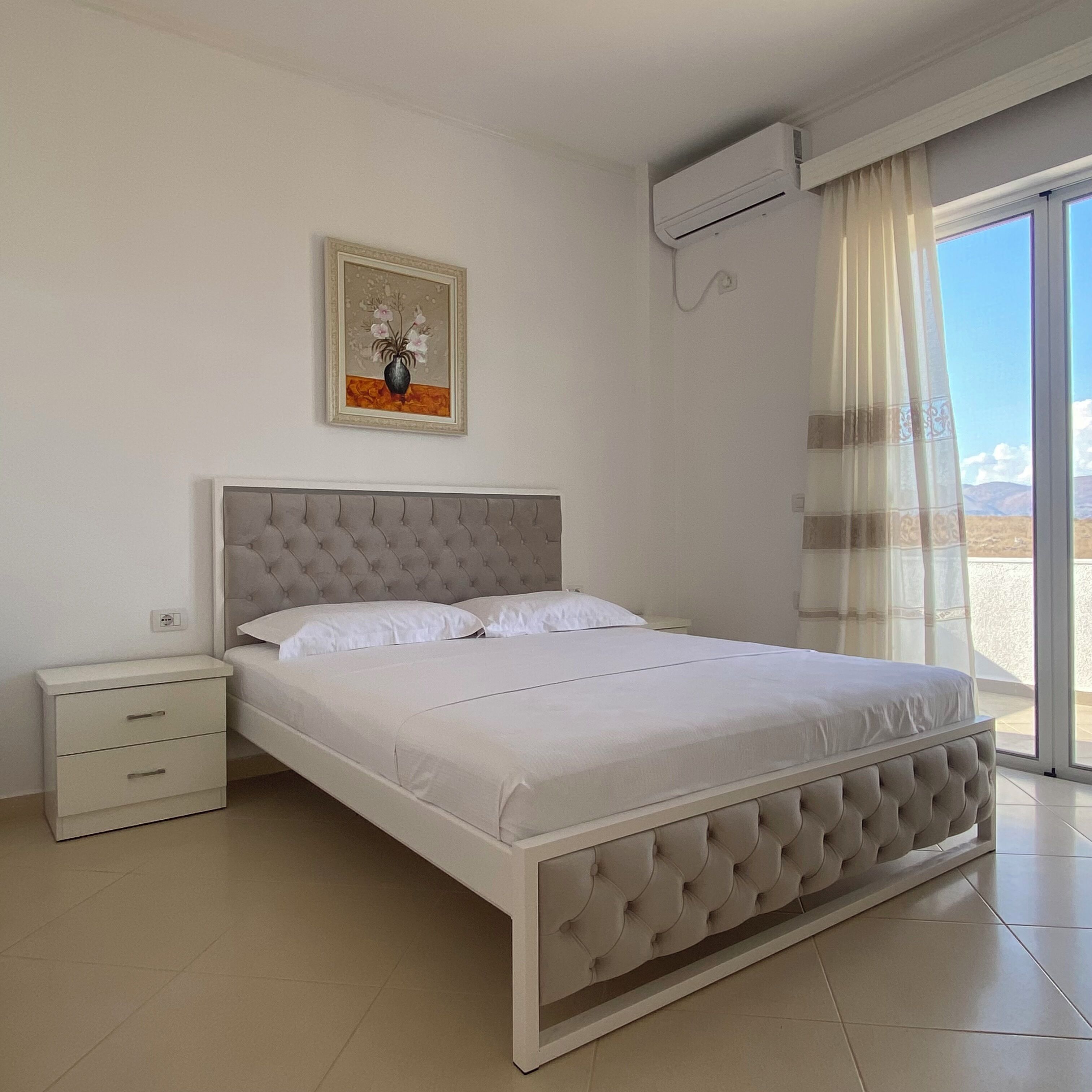 A bedroom with a bed, a nightstand, a wall-mounted air conditioner, and a window with curtains.